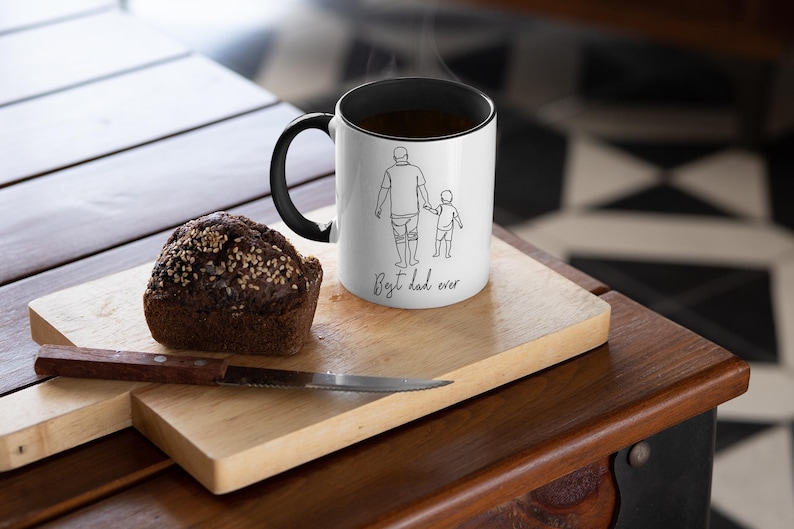 May include: A white coffee mug with a black handle and rim, featuring a line drawing of a parent and child with the text "Best dad ever." A slice of bread and a knife rest on a wooden cutting board.