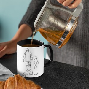May include: A white mug with a black interior and handle is being filled with tea. The mug features a line drawing of a parent and child holding hands, with the text "Best dad ever". A croissant sits in the foreground.