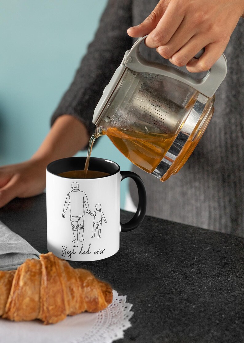 May include: A white mug with a black interior and handle is being filled with tea. The mug features a line drawing of a parent and child holding hands, with the text "Best dad ever". A croissant sits in the foreground.