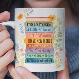 Op de afbeelding: Witte keramische mok met een stapel kleurrijke boeken, waaronder "Pride and Prejudice" en "The Great Gatsby". Bloemenaccenten omringen de boekenstapel. De mok wordt door een persoon vastgehouden.