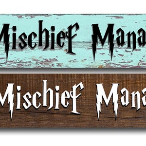 May include: Two rectangular wooden signs with the text "Mischief Manager". One sign is light blue with black text, the other is brown with white text. The signs have a distressed, rustic appearance.