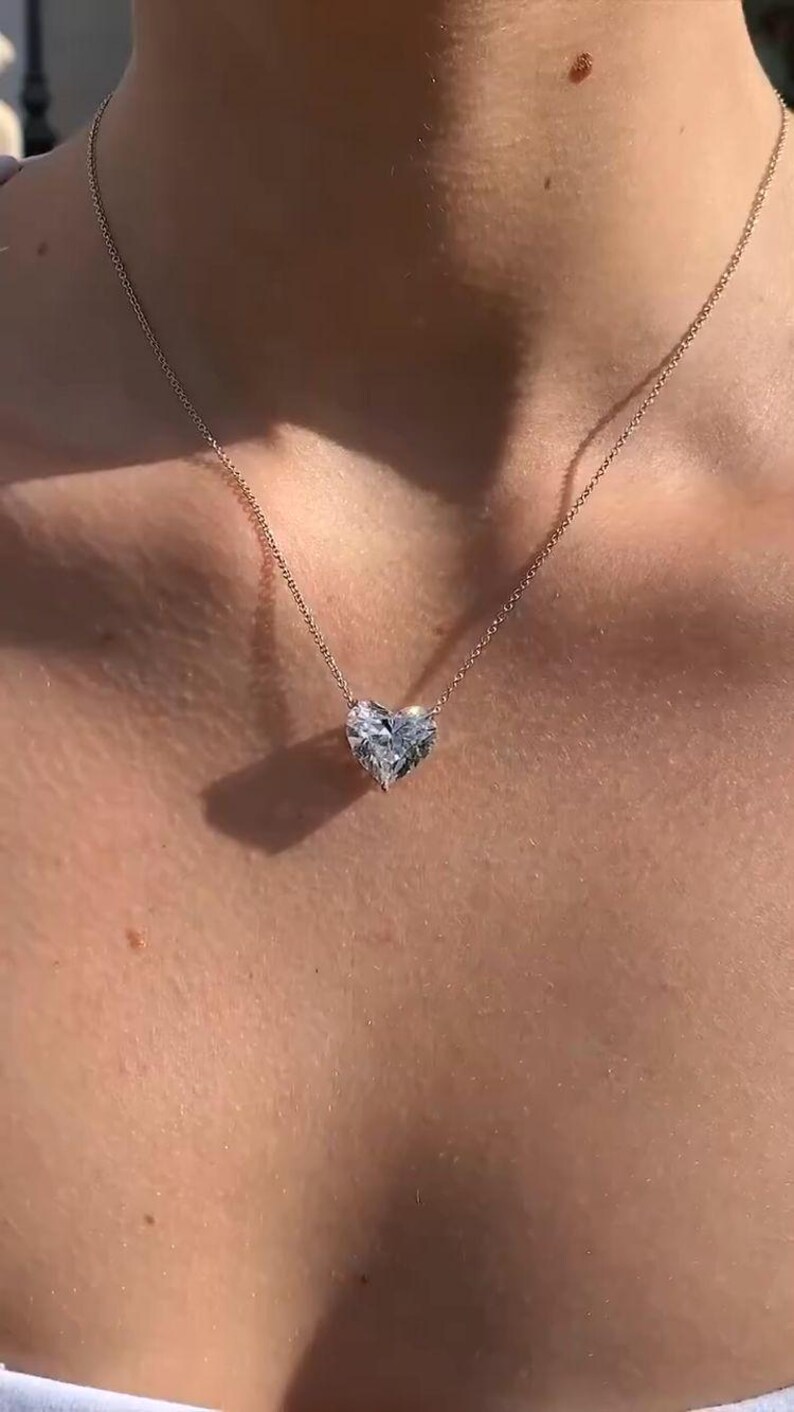 May include: A delicate gold necklace featuring a heart-shaped diamond pendant. The pendant is faceted, catching the light, and suspended from a thin chain. The necklace rests on a person's skin, highlighting the jewelry's design.