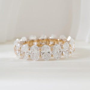 May include: An eternity band ring featuring a continuous row of oval-cut clear gemstones set in a polished yellow gold metal band. The ring displays a series of identical oval stones secured by small gold prongs, creating a uniform circular design.