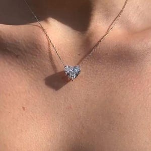 May include: A delicate necklace featuring a heart-shaped pendant. The pendant is made of a clear, sparkling material, possibly a gemstone or crystal. The chain is a thin, silver-colored metal, and the necklace rests on a person's skin.