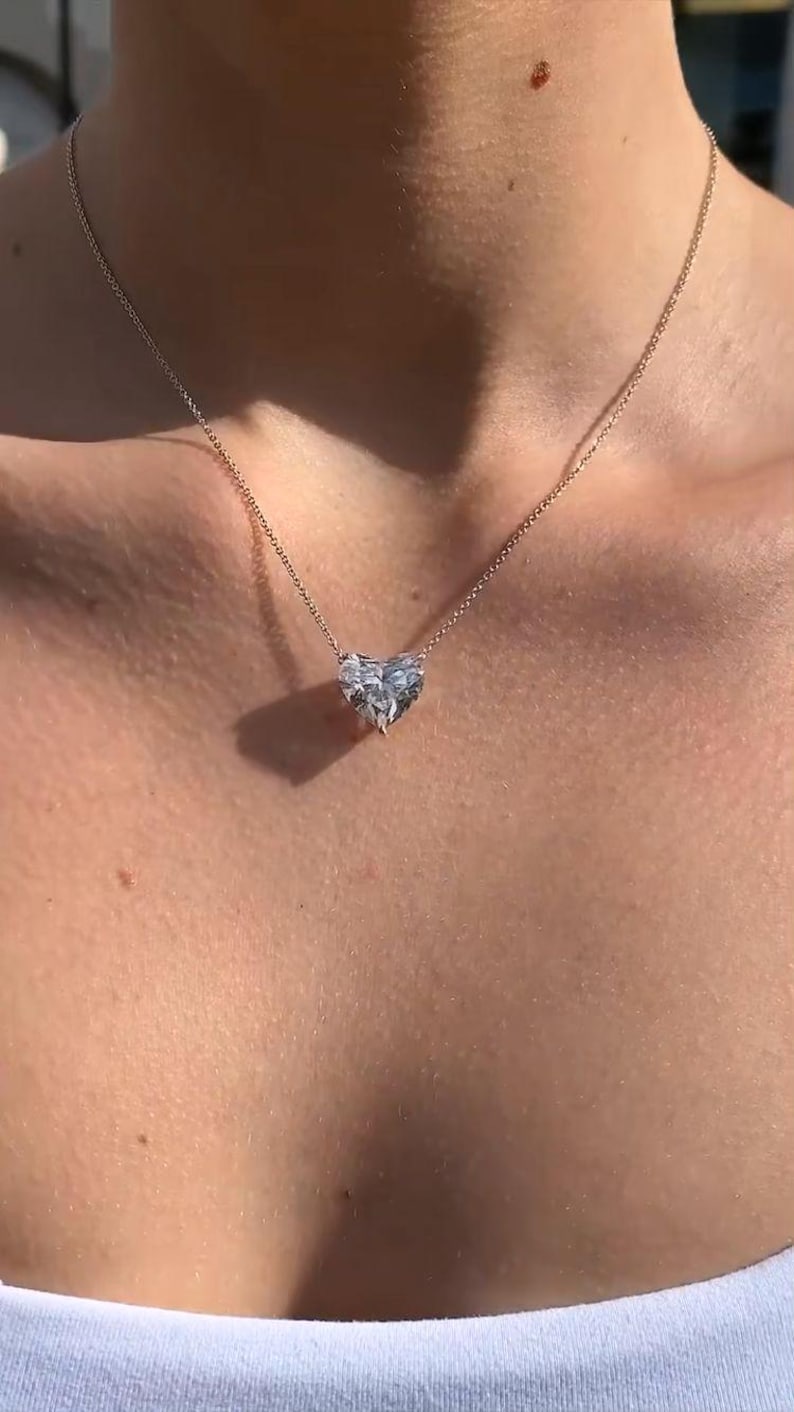 May include: A delicate necklace featuring a heart-shaped pendant. The pendant is made of a clear, sparkling material, possibly a gemstone or crystal. The chain is a thin, silver-colored metal, and the necklace rests on a person's skin.