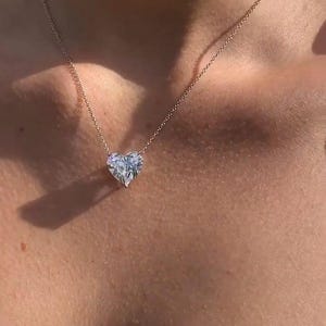 May include: A delicate gold necklace featuring a heart-shaped diamond pendant. The pendant is a clear, faceted stone, suspended from a fine chain. The necklace is worn on a person's neck, showcasing the jewelry's design.