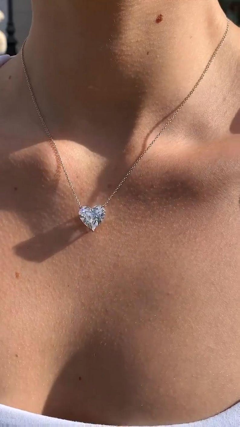 May include: A delicate gold necklace featuring a heart-shaped diamond pendant. The pendant is a clear, faceted stone, suspended from a fine chain. The necklace is worn on a person's neck, showcasing the jewelry's design.
