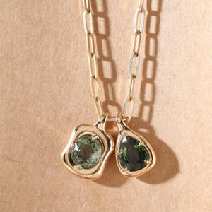 May include: A gold-toned necklace featuring two pendants with green gemstones. The pendants have unique, irregular shapes and are set in gold bezels. The chain is a linked design, and the background is a neutral tan color.