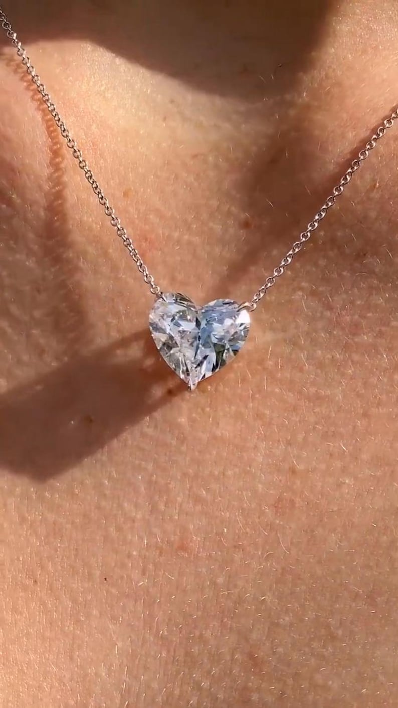 May include: A heart-shaped diamond pendant necklace. The large, clear diamond is suspended from a delicate silver chain. The necklace is displayed against a skin tone background, highlighting the jewelry's sparkle and design.