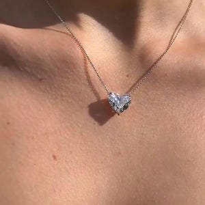 May include: A delicate necklace featuring a heart-shaped pendant. The pendant is a clear, faceted gemstone, suspended from a thin, gold-colored chain. The necklace rests on a person's skin, highlighting the jewelry's design.