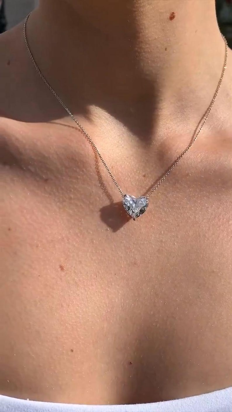 May include: A delicate necklace featuring a heart-shaped pendant. The pendant is a clear, faceted gemstone, suspended from a thin, gold-colored chain. The necklace rests on a person's skin, highlighting the jewelry's design.