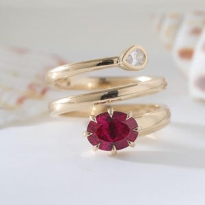Könnte beinhalten: Ein goldener Bypass-Ring mit einem leuchtend oval geschliffenen roten Edelstein, der an einem Ende von Krappen gehalten wird, und einem birnenförmig geschliffenen klaren Edelstein am anderen Ende. Die Ringschiene ist glatt poliert und spiralförmig gewunden.