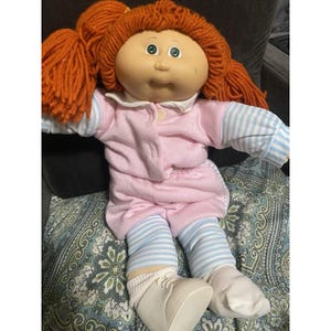 May include: A doll with bright orange yarn hair styled in pigtails, featuring a round face with green eyes. The doll wears a pink long-sleeved top with a white collar, paired with light blue and white striped leggings and white shoes.