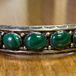 May include: Silver cuff bracelet featuring five oval, vibrant green malachite stones. The bracelet has a detailed, engraved design along the edges. The malachite stones display unique swirling patterns, adding to the bracelet's appeal.