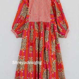 K&ouml;nnte beinhalten: Ein langes Kleid mit einem auff&auml;lligen Tigerstreifenmuster in Orange, Rosa und Beige. Das Oberteil hat vertikale rot-wei&szlig;e Streifen. Das Kleid hat lange &Auml;rmel und h&auml;ngt an einem Holzkleiderb&uuml;gel. Der Name "Shreyadesigng" ist aufgedruckt.