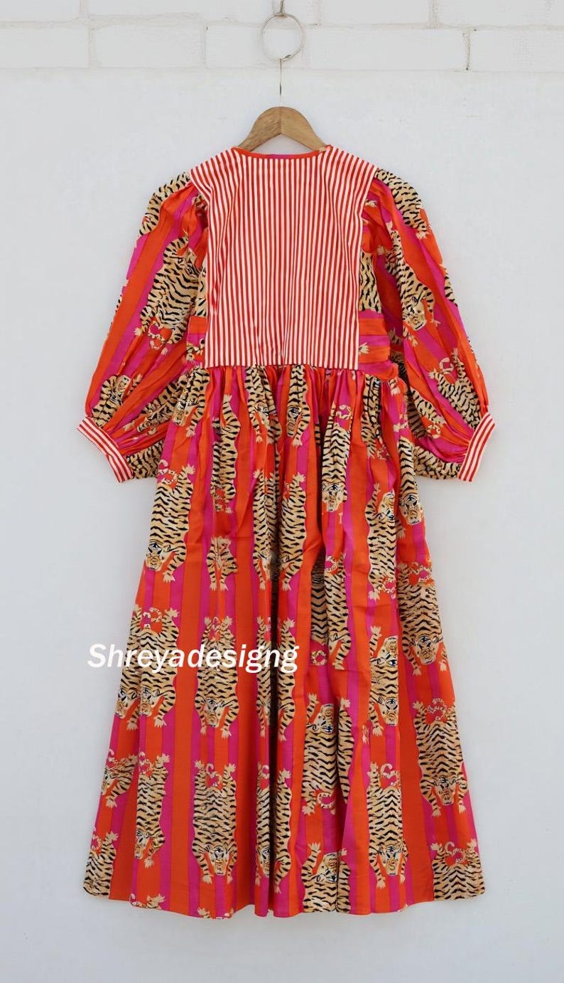 K&ouml;nnte beinhalten: Ein langes Kleid mit einem auff&auml;lligen Tigerstreifenmuster in Orange, Rosa und Beige. Das Oberteil hat vertikale rot-wei&szlig;e Streifen. Das Kleid hat lange &Auml;rmel und h&auml;ngt an einem Holzkleiderb&uuml;gel. Der Name "Shreyadesigng" ist aufgedruckt.