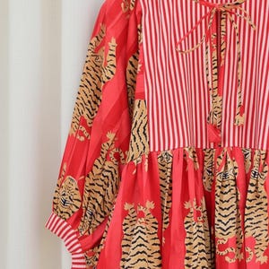 K&ouml;nnte beinhalten: Rot-wei&szlig; gestreiftes Kleid mit Tigerprint. Das Kleid hat lange &Auml;rmel mit gestreiften Manschetten und einen Bindekragen. Der Rock hat ein auff&auml;lliges Tigermuster. Das Kleid h&auml;ngt an einem Holzkleiderb&uuml;gel.