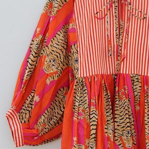 K&ouml;nnte beinhalten: Ein lebendiges Kleid mit einer Mischung aus Mustern. Das Oberteil hat vertikale rot-wei&szlig;e Streifen, w&auml;hrend die &Auml;rmel und der Rock ein Tigerstreifen-Design in Orange, Pink und Schwarz aufweisen. Das Kleid hat eine Schleife am Ausschnitt und den Text "Shreyadesigng".
