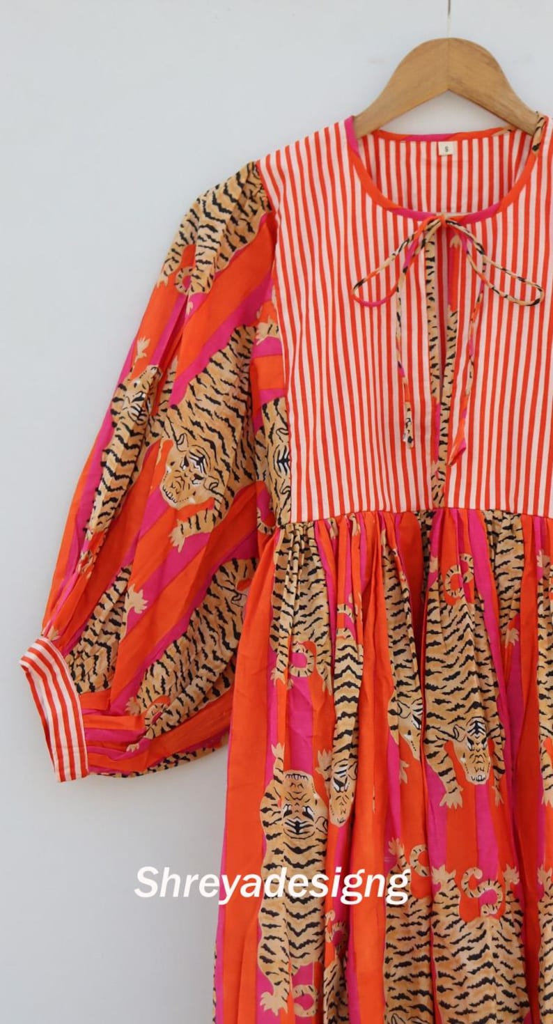 K&ouml;nnte beinhalten: Ein lebendiges Kleid mit einer Mischung aus Mustern. Das Oberteil hat vertikale rot-wei&szlig;e Streifen, w&auml;hrend die &Auml;rmel und der Rock ein Tigerstreifen-Design in Orange, Pink und Schwarz aufweisen. Das Kleid hat eine Schleife am Ausschnitt und den Text "Shreyadesigng".