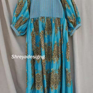 K&ouml;nnte beinhalten: Ein langes Kleid mit einem blau-wei&szlig; gestreiften Oberteil und &Auml;rmeln. Der Rock hat ein sich wiederholendes Tigerstreifenmuster in Beige- und Schwarzt&ouml;nen. Der Text "Shreyadesigng" ist auf der Vorderseite des Kleides zu sehen.