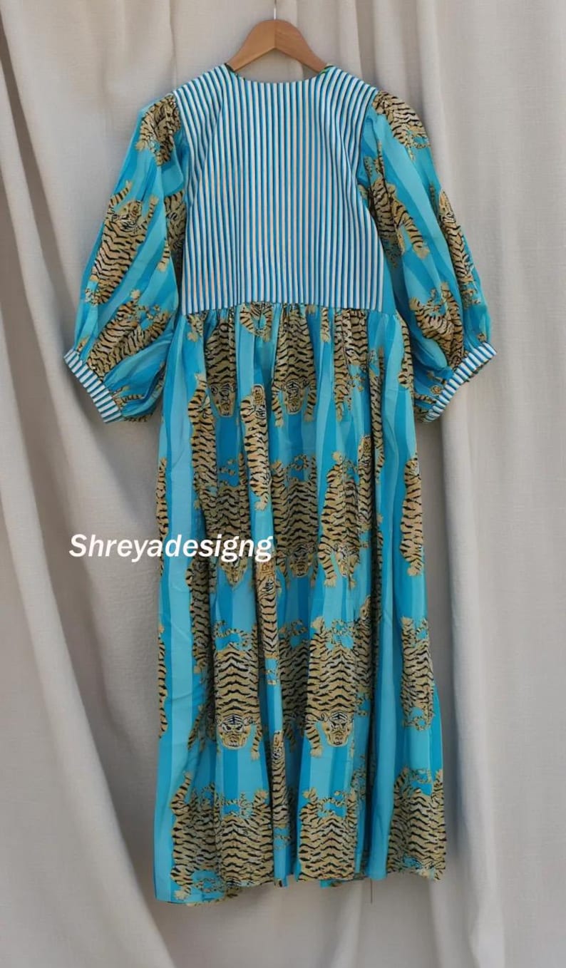 K&ouml;nnte beinhalten: Ein langes Kleid mit einem blau-wei&szlig; gestreiften Oberteil und &Auml;rmeln. Der Rock hat ein sich wiederholendes Tigerstreifenmuster in Beige- und Schwarzt&ouml;nen. Der Text "Shreyadesigng" ist auf der Vorderseite des Kleides zu sehen.