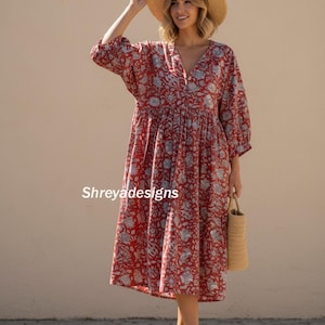 Könnte beinhalten: Rotes Midikleid mit floralem Muster, V-Ausschnitt und Dreiviertelärmeln. Das Kleid hat eine geraffte Taille und wird mit einem Strohhut und einer gewebten Handtasche getragen. Der Text "Shreyadesigns" ist auf das Kleid gedruckt.