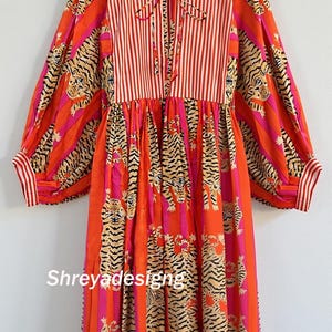 K&ouml;nnte beinhalten: Langes Kleid mit Tigerstreifenmuster in Orange, Pink und Schwarz. Das Oberteil hat rot-wei&szlig;e vertikale Streifen und einen Bindekragen. Lange &Auml;rmel mit Manschetten. Der Name "Shreyadesigng" ist sichtbar.