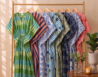 May include: A collection of eight kaftans hanging on a wooden rack. The garments feature various patterns including green and yellow plaid, pink and navy stripes, blue floral prints, and red stripes. Each kaftan has a V-neck and a drawstring waist.