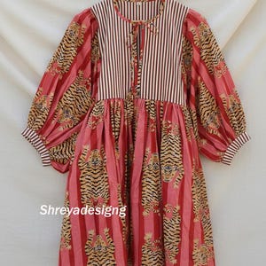 K&ouml;nnte beinhalten: Ein lang&auml;rmliges Kleid mit gestreiftem Oberteil und einem Rock mit Tigerprint. Das Kleid hat einen runden Ausschnitt mit Krawattenverschluss und volumin&ouml;sen &Auml;rmeln. Die Farbpalette umfasst Rot, Rosa und Beige.