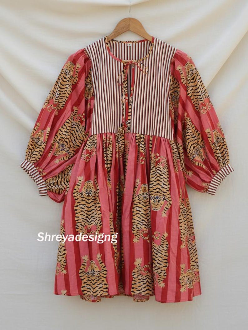 K&ouml;nnte beinhalten: Ein lang&auml;rmliges Kleid mit gestreiftem Oberteil und einem Rock mit Tigerprint. Das Kleid hat einen runden Ausschnitt mit Krawattenverschluss und volumin&ouml;sen &Auml;rmeln. Die Farbpalette umfasst Rot, Rosa und Beige.