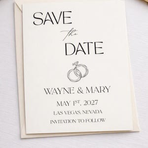 Könnte beinhalten: Eine cremefarbene Save-the-Date-Karte mit schwarzem Text, der SAVE the DATE, WAYNE & MARY, MAY 1ST, 2027, LAS VEGAS, NEVADA, INVITATION TO FOLLOW lautet, sowie eine Strichzeichnung von zwei ineinandergreifenden Ringen mit einem Diamanten.