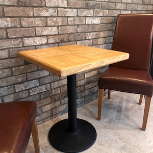 Puede incluir: Una mesa cuadrada de madera con tapa marrón claro y base de metal negro. Una silla de cuero marrón y un taburete a juego están colocados cerca de la mesa. El fondo presenta una pared de ladrillo.
