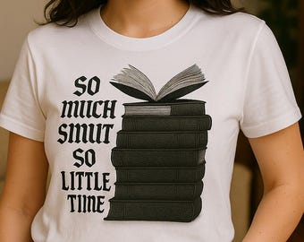 Può includere: T-shirt bianca con la scritta "SO MUCH SMUT SO LITTLE TIME" in nero, con un carattere in stile gotico. La grafica presenta una pila di libri neri con un libro aperto sopra. La persona indossa jeans blu.