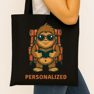 Op de afbeelding: Zwarte draagtas met een cartoon Bigfoot met een zonnebril en een rugzak, die een boek leest. De Bigfoot is bruin met oranje accenten. Het woord "PERSONALIZED" staat in het oranje onderaan de tas.