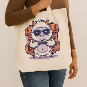 May include: Cream-colored tote bag featuring a cartoon yeti wearing sunglasses and a backpack, reading a book. The yeti is white with orange horns and a purple belly. The backpack is orange.