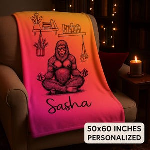 May include: A personalized blanket with a gradient of orange, pink, and red. It features a black illustration of a meditating Bigfoot, the name "Sasha", and the text "50x60 INCHES PERSONALIZED". The blanket is draped over a chair.
