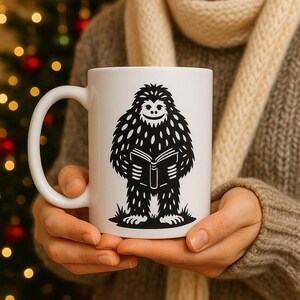 May include: White ceramic mug with a black and white illustration of a Bigfoot reading a book. The Bigfoot has a smiling face and is standing on grass. The mug has a large handle. The mug is held by a person.