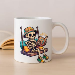 Puede incluir: Taza de cerámica blanca con un dibujo de un esqueleto leyendo un libro en un columpio. El esqueleto lleva un sombrero colorido y zapatillas verdes. El libro tiene un efecto brillante.