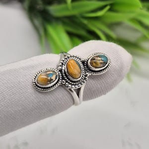 Puede incluir: Anillo de plata con tres piedras ovales. La piedra central es naranja, mientras que las piedras laterales tienen tonos azules y naranjas. El anillo tiene un engaste detallado y ornamentado.