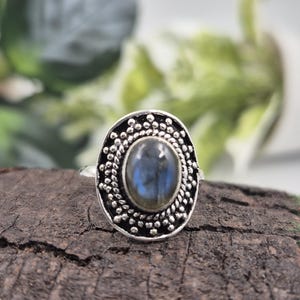 Puede incluir: Un anillo de plata con una piedra preciosa ovalada de color azul grisáceo. La piedra preciosa está engastada en un bisel decorativo con cuentas. El anillo se muestra sobre un trozo de madera oscura y texturizada, con follaje verde al fondo.