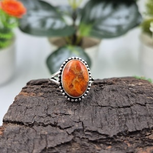 Puede incluir: Anillo de plata con una piedra ovalada naranja con un patrón moteado. La piedra está engastada en un bisel con cuentas. El anillo se muestra sobre un trozo de madera oscura y texturizada, con vegetación borrosa al fondo.