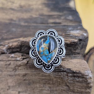 Puede incluir: Anillo plateado con una piedra en forma de lágrima. La piedra presenta tonos azules y marrones, engastada en un bisel decorativo y festoneado. El anillo se muestra sobre una superficie de madera rústica.