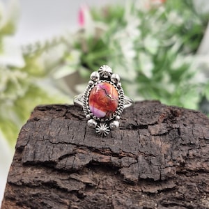Puede incluir: Un anillo de plata con una piedra ovalada multicolor. La piedra presenta tonos de rojo, naranja y morado. El anillo tiene un engaste detallado y ornamentado con pequeños elementos decorativos.