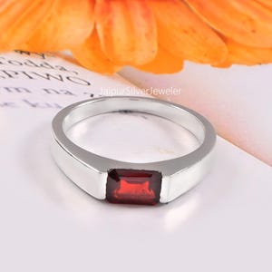 May include: A silver ring with a rectangular, deep red gemstone. The ring is set against a white background with an orange flower in the background. The ring is a simple, elegant design.