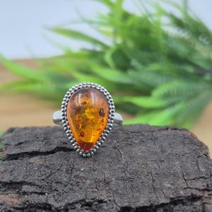 Puede incluir: Un anillo de plata con una piedra de ámbar en forma de lágrima. El ámbar es de color naranja cálido con inclusiones visibles. El anillo está sobre una superficie oscura y texturizada, con follaje verde en el fondo. El engaste del anillo tiene un borde de cuentas.