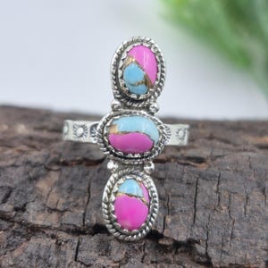 Puede incluir: Anillo de plata con tres piedras ovaladas. Cada piedra está dividida en secciones rosa y azul claro, con un borde plateado en forma de cuerda. Se encuentra sobre una superficie de madera oscura.