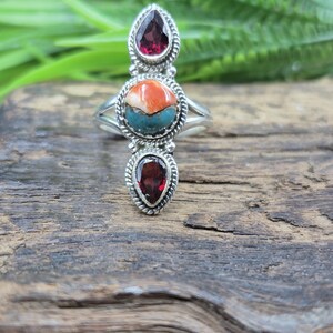 Puede incluir: Un anillo de plata con tres piedras. Las piedras superior e inferior tienen forma de lágrima, de color rojo intenso, y están engastadas en un borde en forma de cuerda. La piedra central es redonda, con colores naranja, turquesa y blanco. El anillo se muestra sobre una superficie de madera.