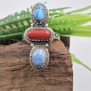 Puede incluir: Un anillo de plata con tres piedras: una piedra roja rectangular central y dos piedras azules ovaladas. El anillo tiene un borde detallado en forma de cuerda alrededor de cada piedra. El anillo está sobre una superficie de madera.