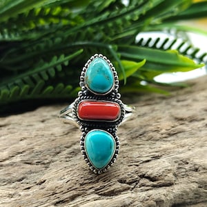 Puede incluir: Un anillo de plata con tres piedras: dos turquesas en forma de lágrima y una piedra rectangular de coral rojo. El anillo está sobre una superficie de madera, con follaje verde de fondo. El diseño del anillo es detallado y ornamentado.