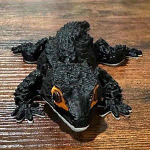 May include: A black lizard figurine with textured skin and orange eyes. The reptile has four legs with white tips on the toes. The figurine is lying on a wooden surface.